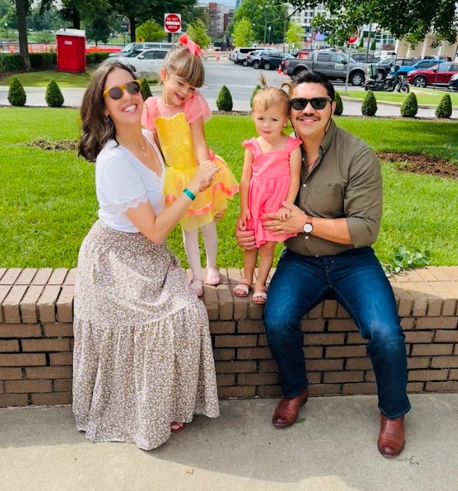 A man and woman sit on a low wall smiling while holding two young girls.