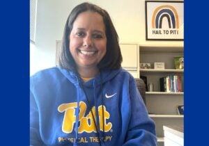 A woman with medium length brown hair wears a blue sweatshirt that says Pitt Physical Therapy