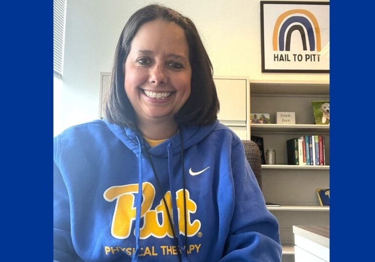 A woman with medium length brown hair wears a blue sweatshirt that says Pitt Physical Therapy