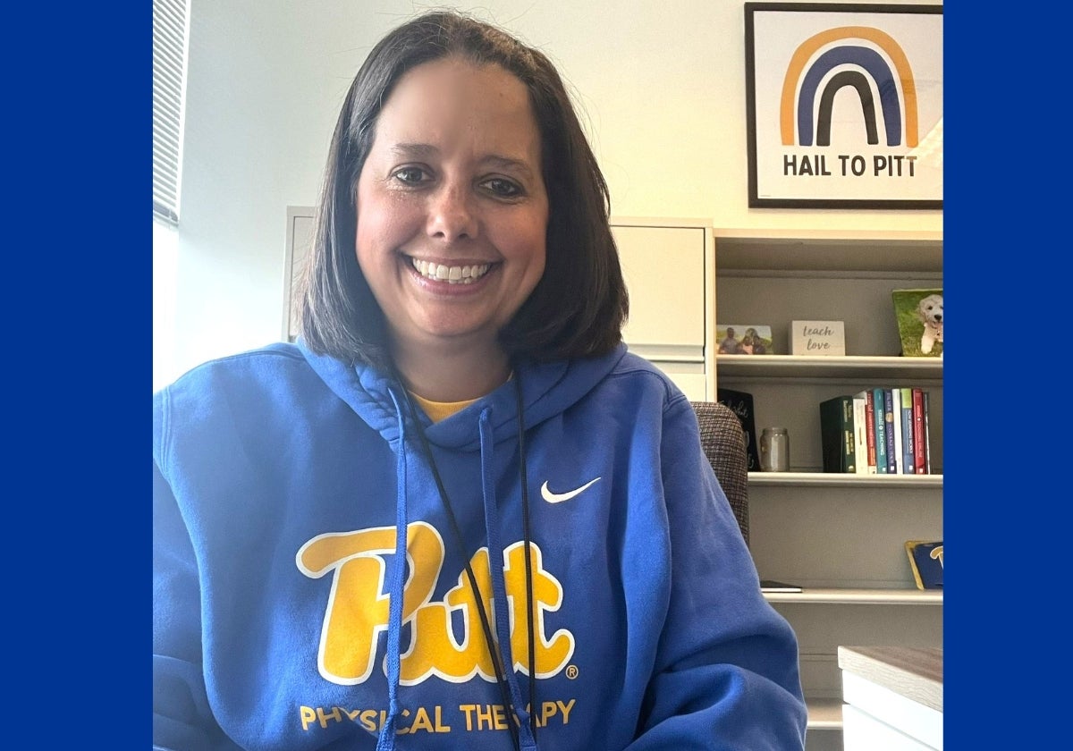 A woman with medium length brown hair wears a blue sweatshirt that says Pitt Physical Therapy