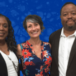 A Black woman with long curly hair stands next to a white woman with short dark gray hair who stands next to a Black man with short dark hair and mustache