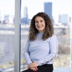 A woman with long curly brown hair wears a lilac colored blouse posing for a photo with the city in the background