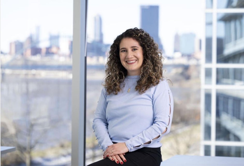 A woman with long curly brown hair wears a lilac colored blouse posing for a photo with the city in the background