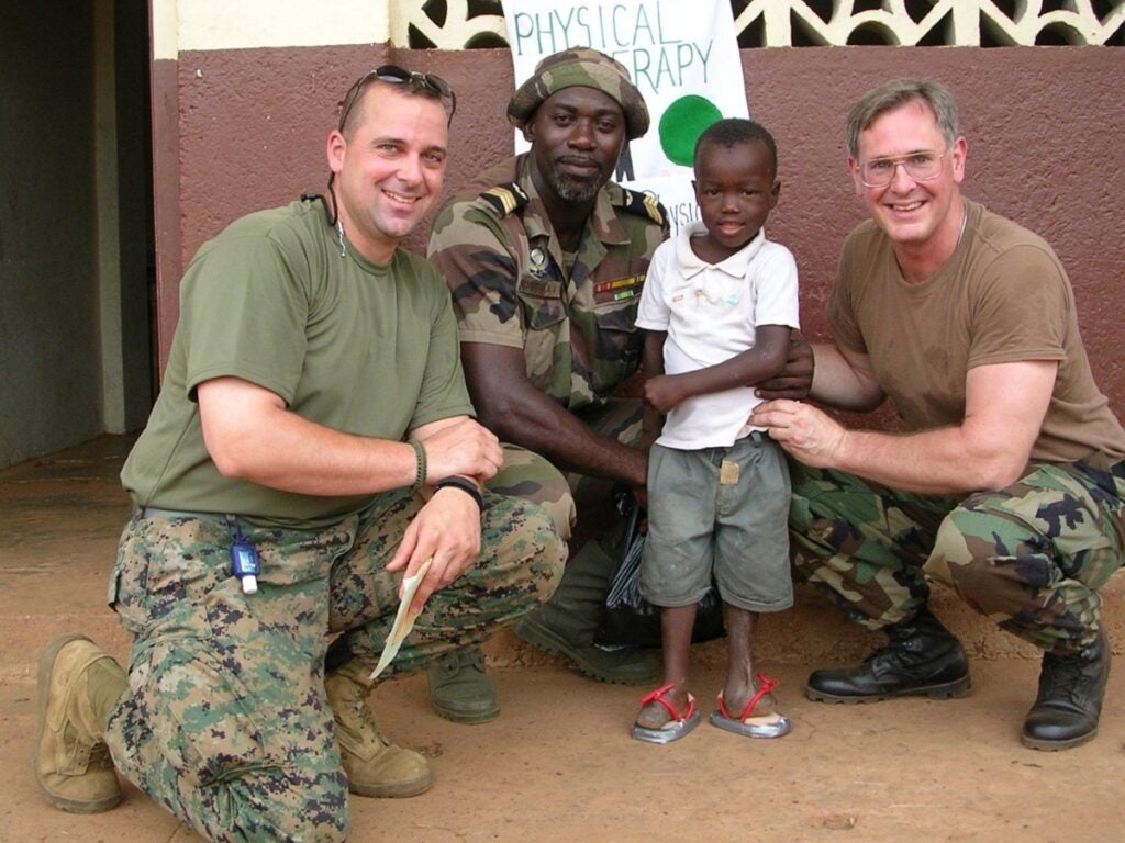 Three men in military fatigues kneel and smile with a young boy wearing sandals on his amputated feet.