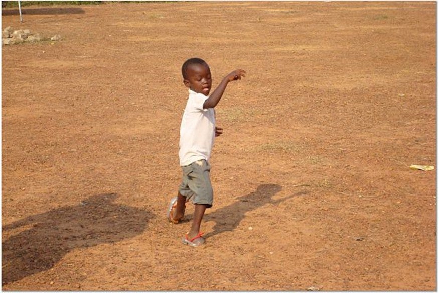 A young boy with short black hair and dark brown skin dances in a field of brown dirt