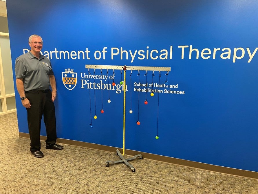 A man with gray hair and glasses stands in front of a blue wall and next to a measurement device with colorful balls hanging from its top