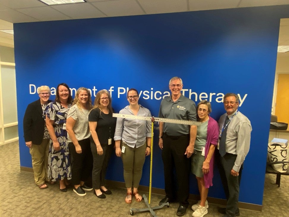 A group of eight people stand in front of a blue wall with a measuring device in front of them