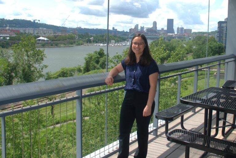 A woman with long brown hair and glasses smiles in front of the city of Pittsburgh skyline
