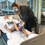 A woman in a green sweatshirt wearing a mask moves the leg of a manikin laying in a hospital bed.
