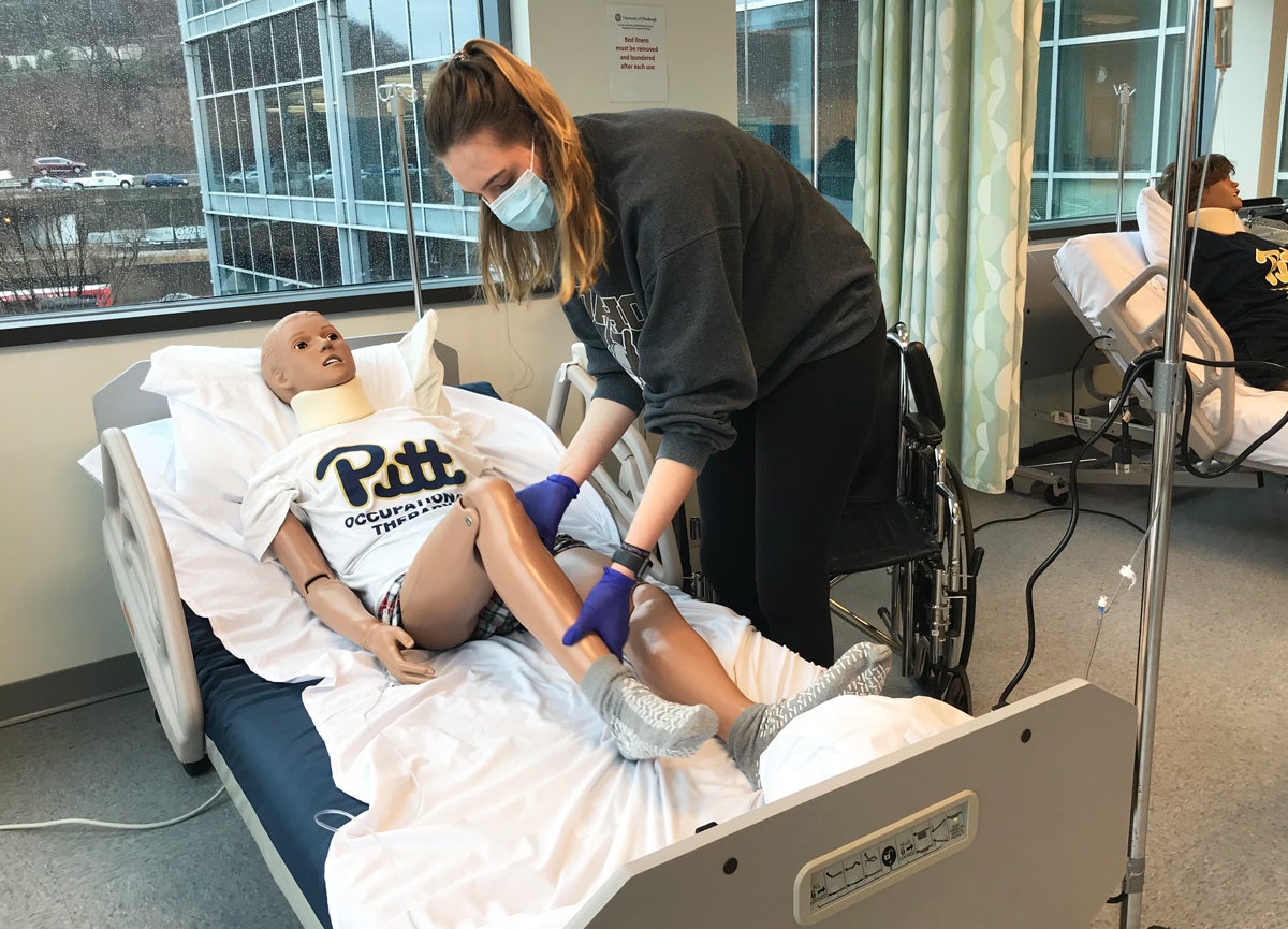 A woman in a green sweatshirt wearing a mask moves the leg of a manikin laying in a hospital bed.