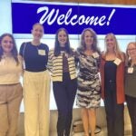 A group of six women wearing conference badges pose for the camera in front of a digital welcome sign