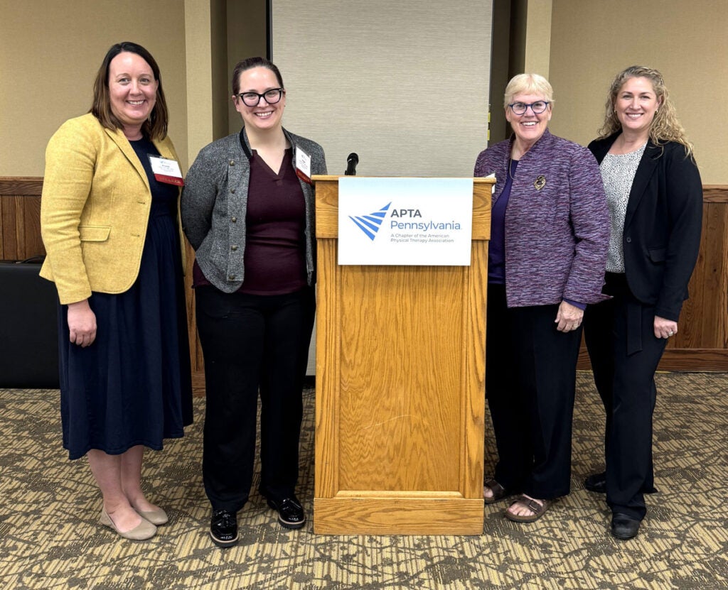 Four women stand beside a conference podium with a sign that says APTA Pennsylvania