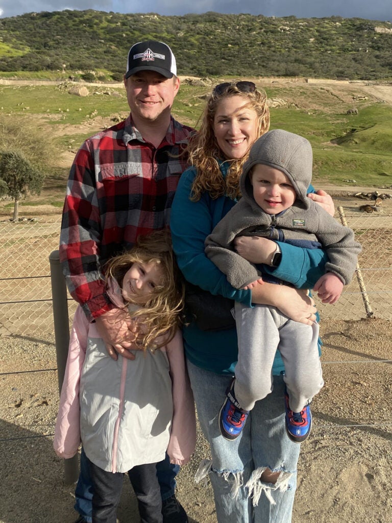 A man and woman stand on a beach with two young children, one in front of the man and the other being held by the woman