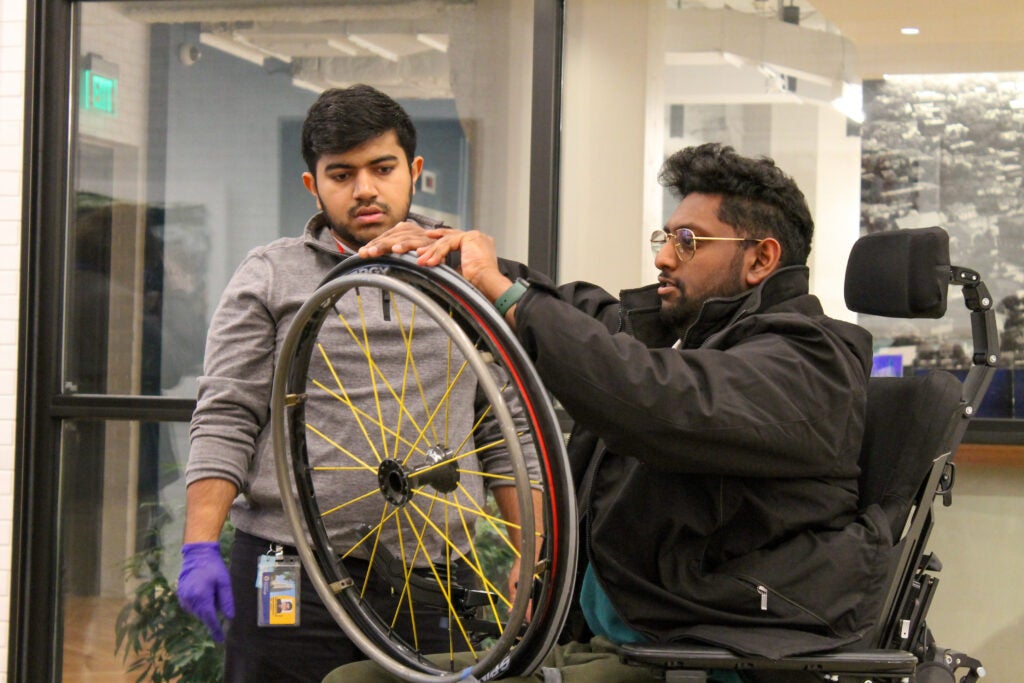 A man with medium skin tone and dark hair sits in a wheelchair while fixing a wheel in his lap