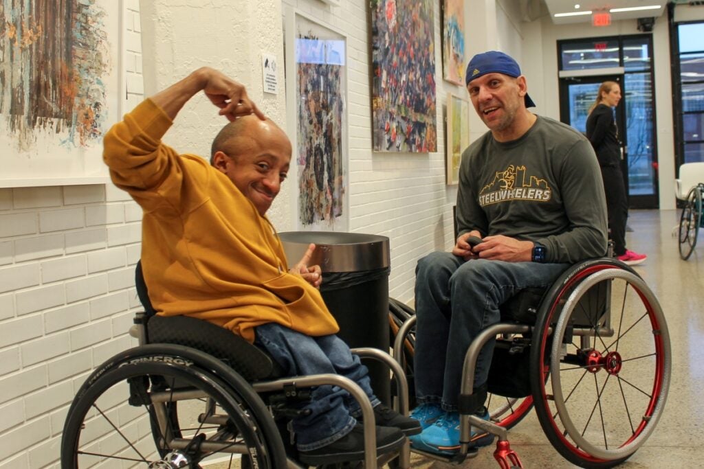 A man with medium skin tone sitting in a wheelchair makes a gesture with his arms while another man in a wheelchair looks on