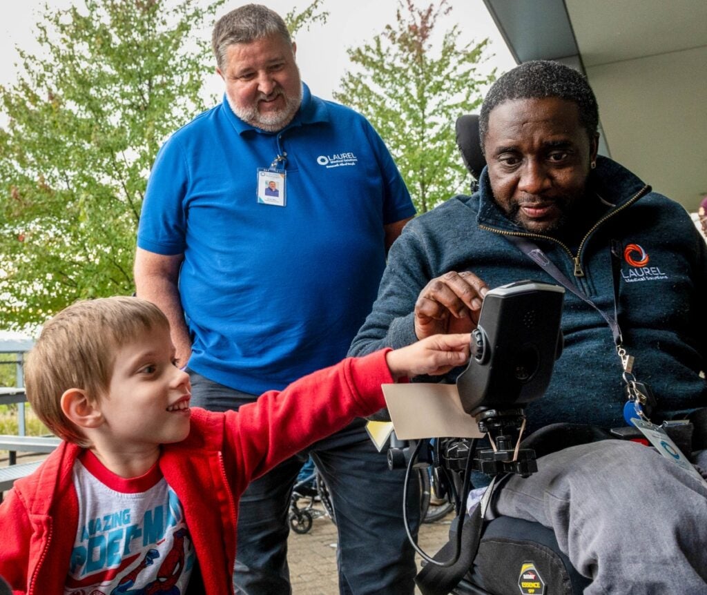A young boy and a man with dark skin tone point at a control panel on the man's wheelchair