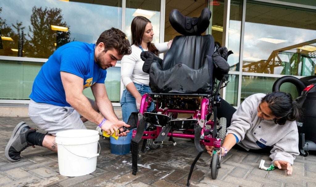 Three students kneel on the ground while washing a wheelchair