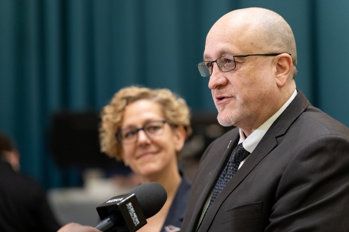 A woman with short curly blonde hair looks on as a man with a gray goatee and glasses speaks to a microphone