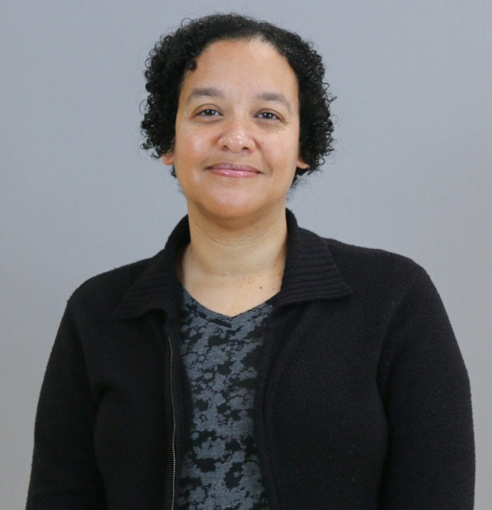 A woman with light brown skin and short dark curly hair wears a black jacket over a gray and black top