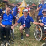 A man wearing a blue team Italia jersey sits in a wheelchair next to a man kneeling wearing a blue polo shirt next to a woman also wearing a blue team Italia jersey sitting in a wheelchair.