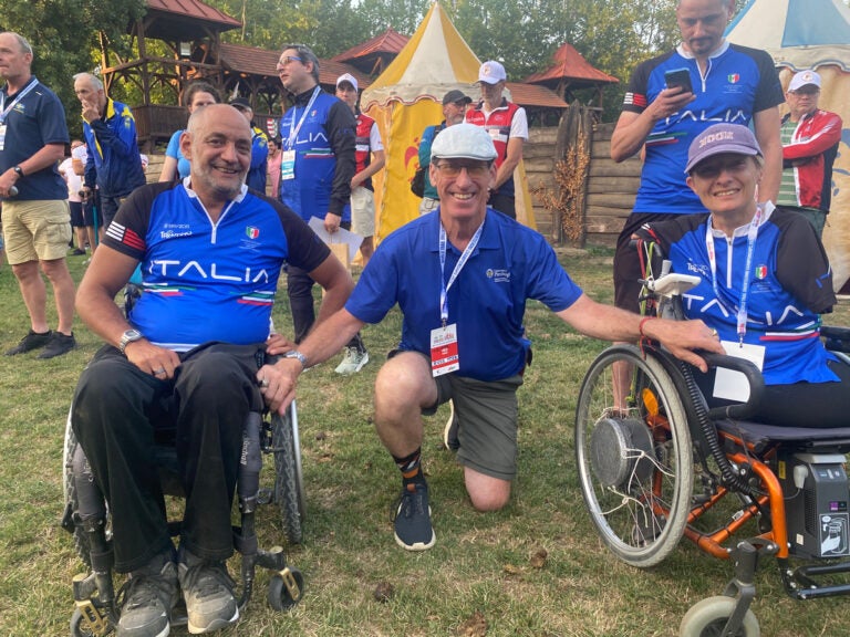 A man wearing a blue team Italia jersey sits in a wheelchair next to a man kneeling wearing a blue polo shirt next to a woman also wearing a blue team Italia jersey sitting in a wheelchair.