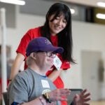 A woman in a red shirt with long black hair smiles at a man with a disability sitting in a chair wearing a gray shirt and ball cap
