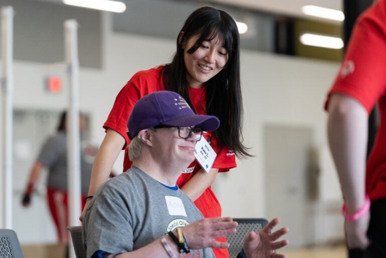 A woman in a red shirt with long black hair smiles at a man with a disability sitting in a chair wearing a gray shirt and ball cap
