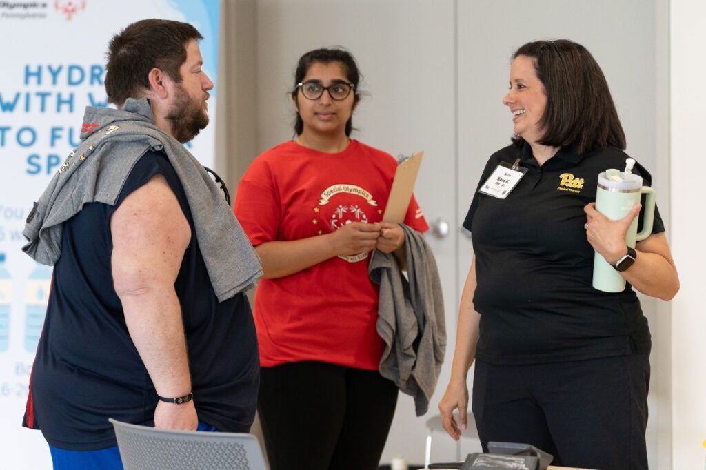A woman with medium length dark hair smiles and talks to a man wearing gym clothes while a woman in a red shirt looks on