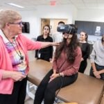 A female professor wearing a pink sweater instructs a student wearing large, specialized goggles while classmates look on