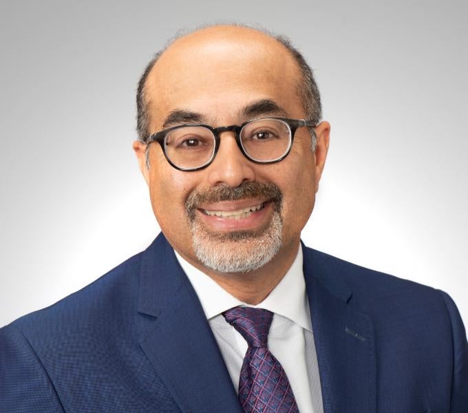 A man with light brown skin and a gray goatee wears glasses and a navy jacket over a white dress shirt and blue tie