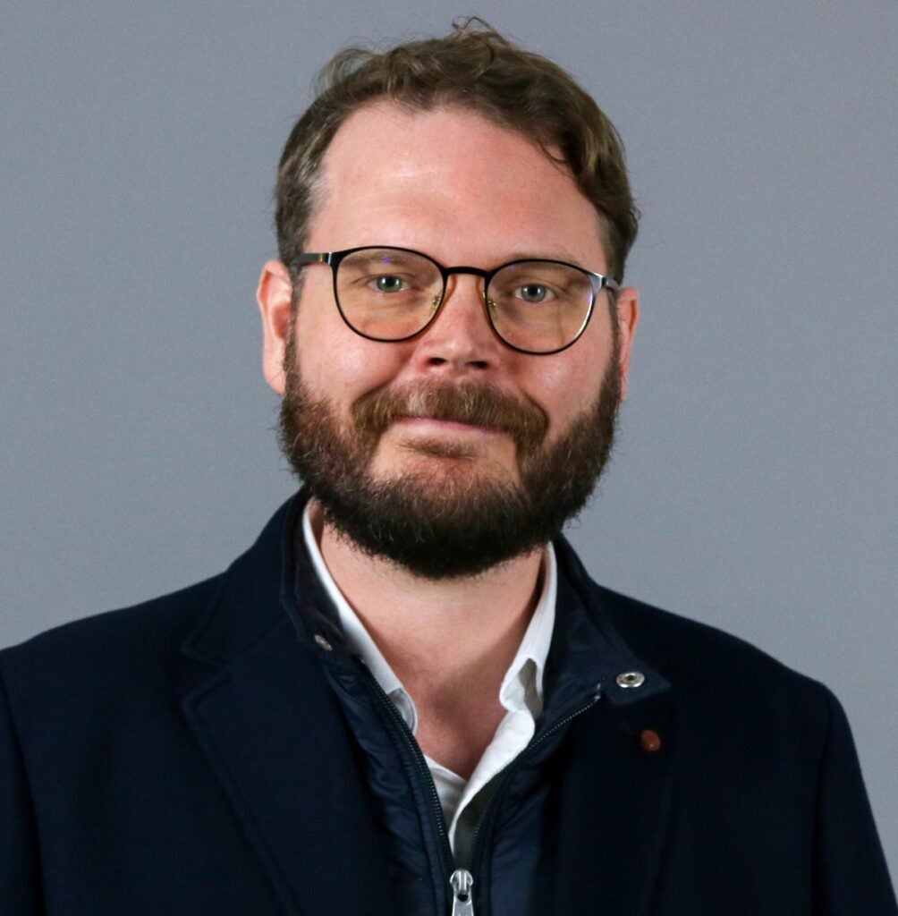 Man with brown hair and brown beard wearing glasses and a dark blue jacket