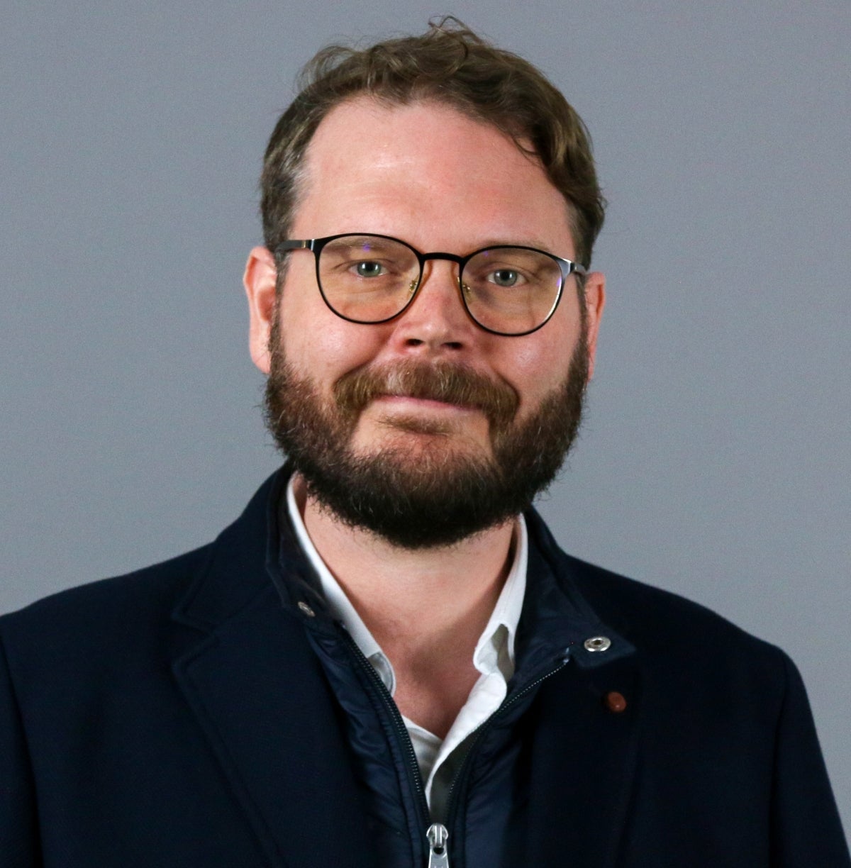 Man with brown hair and brown beard wearing glasses and a dark blue jacket