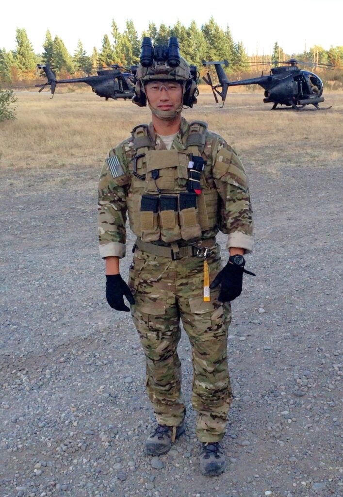 A man is dressed in full Army fatigues and helmet with two black helicopters in the background.