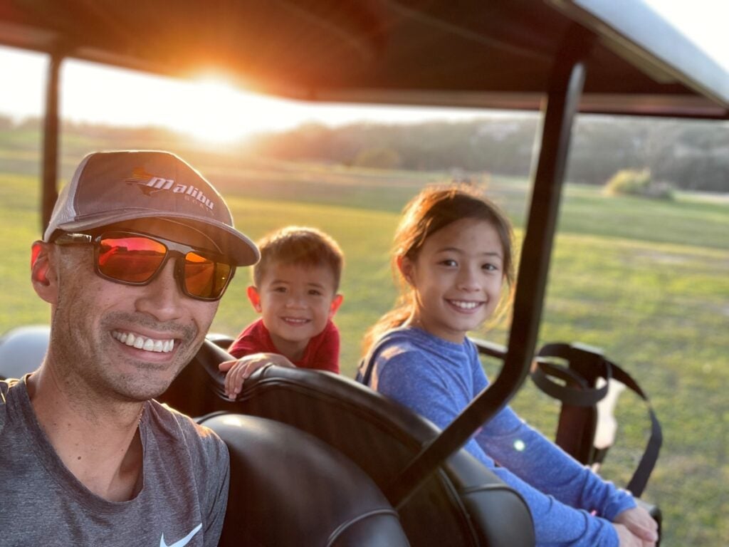 A man in a baseball hat and sunglasses smiles while sitting in a golf cart with a young boy and girl