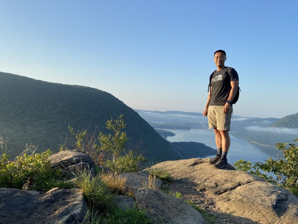 A man in hiking clothes stands on a rock overlooking a valley and lake.