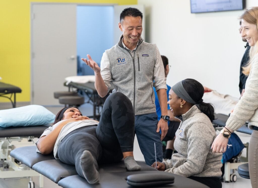 Students sit around an exam table smiling while an instructor stands smiling at them