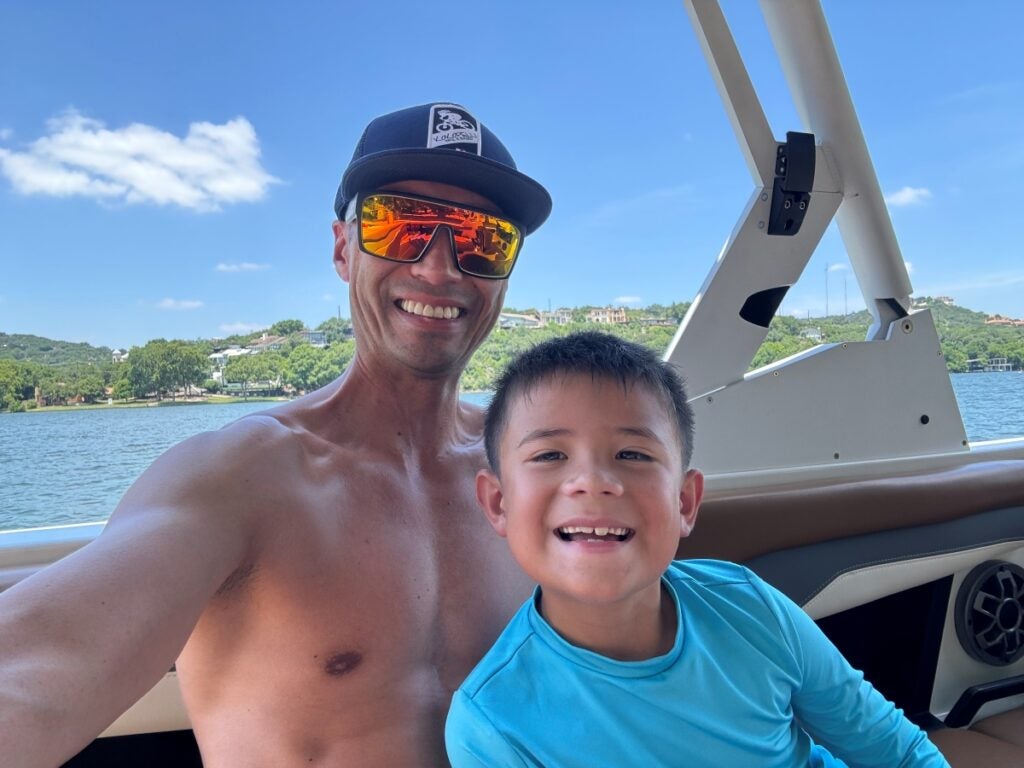 A man in a baseball cap and sunglasses smiles next to a young boy while sitting in a boat on a lake