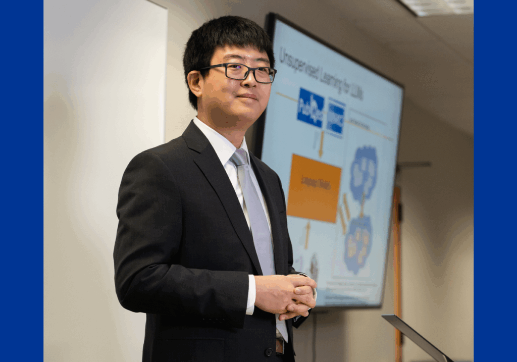 A man with Asian features wearing glasses and a black suit jacket and silver tie poses in front of a large digital screen.