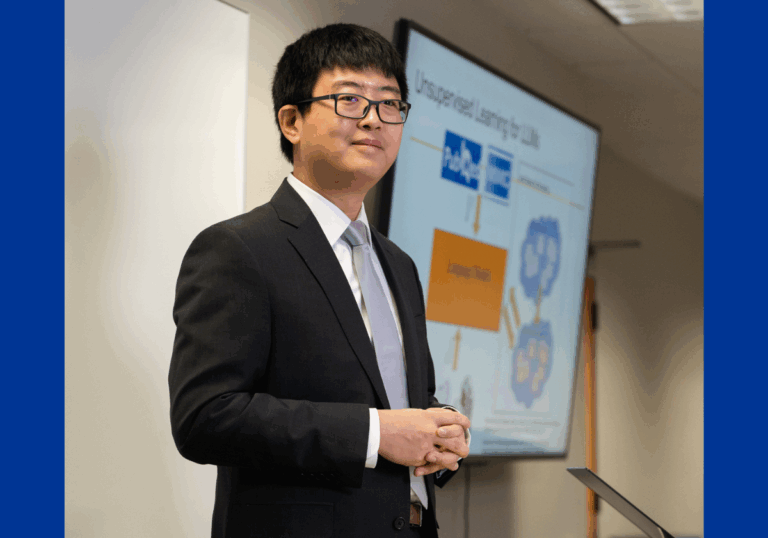 A man with Asian features wearing glasses and a black suit jacket and silver tie poses in front of a large digital screen.