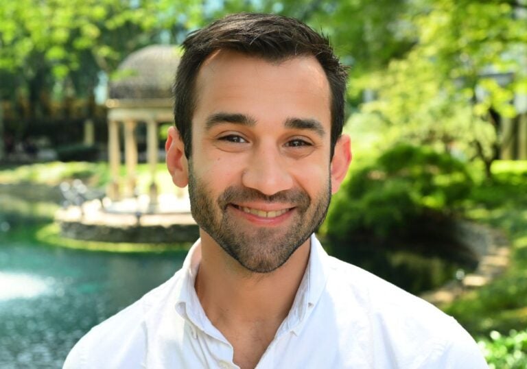 A man with short dark hair and a beard wearing a white collared shirt in front of a green scenic background.