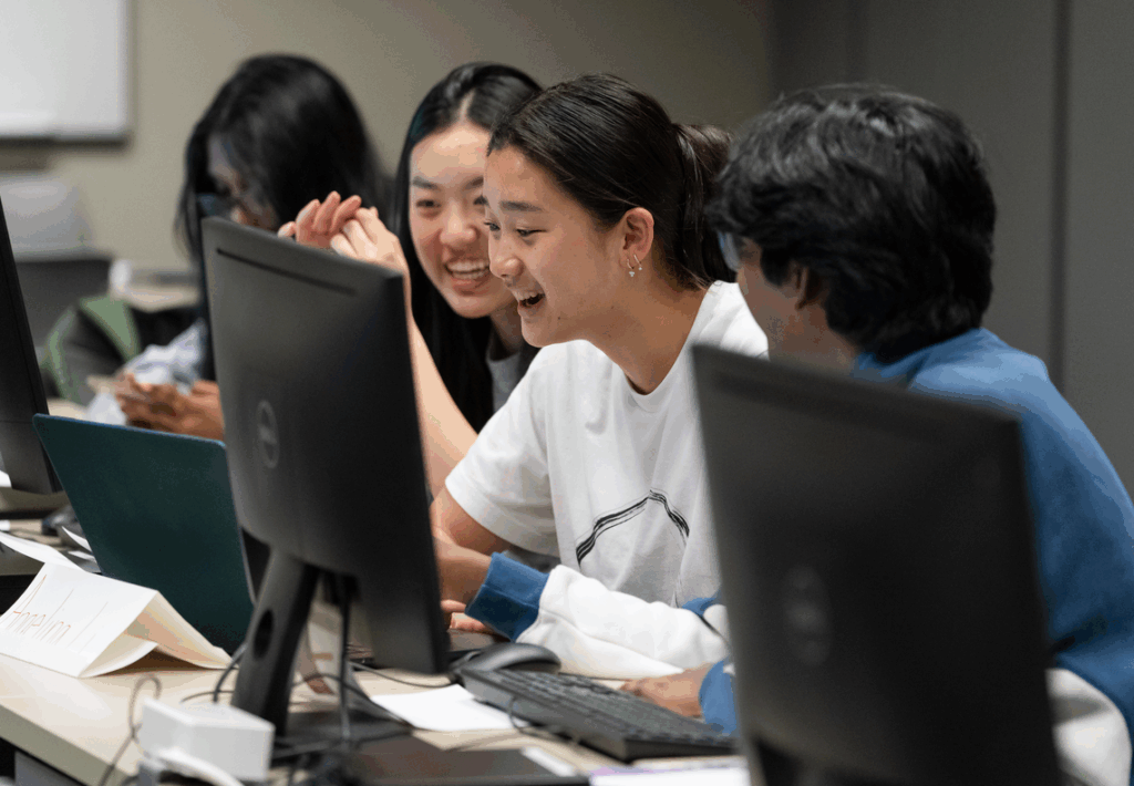 A small group of young men and women sitting together at a desk laughing while looking at a computer.
