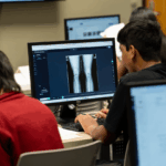 A young man with dark hair wearing a black t-shirt sits at a table in front of a computer where an image is displayed of an x-ray of a person's legs.