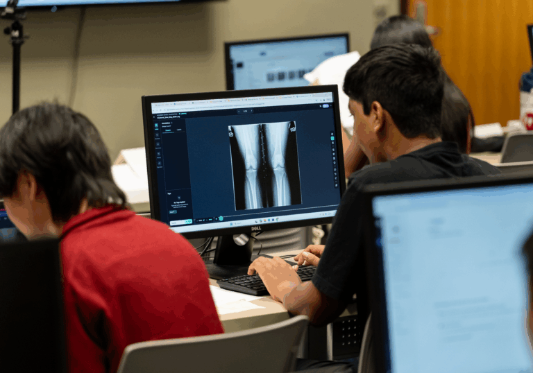 A young man with dark hair wearing a black t-shirt sits at a table in front of a computer where an image is displayed of an x-ray of a person's legs.