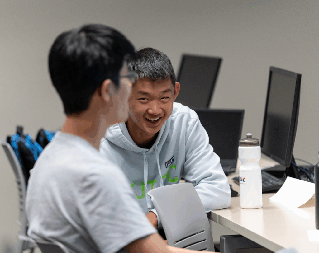 A young man with short hair wearing a light blue sweatshirt smiles at a young man with dark hair and a gray shirt who he is sitting next to at a desk with computers.