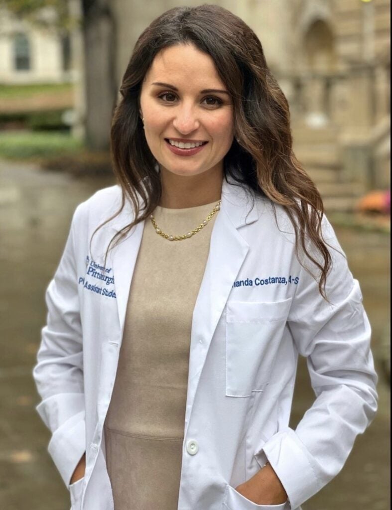 Woman with brown hair wearing a white lab coat over a tan dress and a gold necklace.