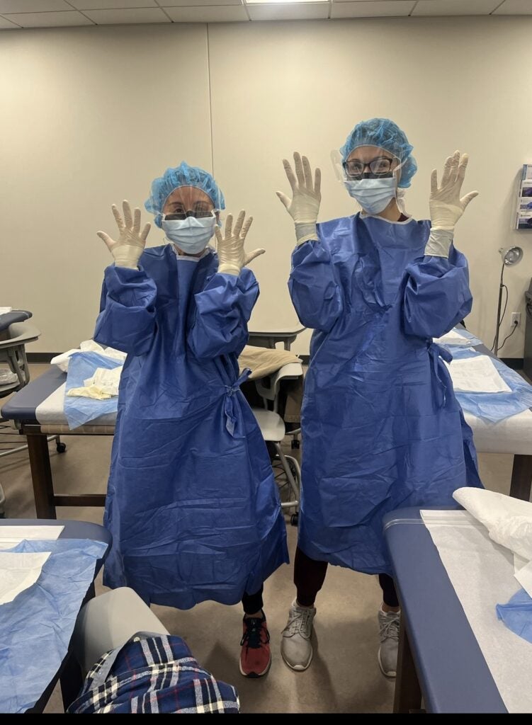 Two women wearing blue scrubs, hairnets and white gloves and face masks in a clinical setting.