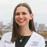 Woman with straight brown hair wearing a white lab coat over a black dress with a silver Star of David necklace and silver earrings.