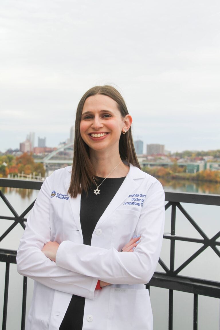 A woman with mid-length straight brown hair wearing a white lab coat over a black dress and s silver Star of David necklace standing in front of the Pittsburgh skyline.