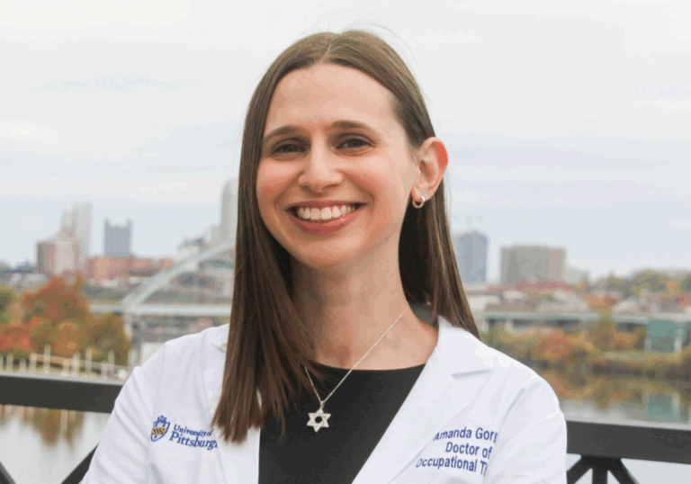 Woman with straight brown hair wearing a white lab coat over a black dress with a silver Star of David necklace and silver earrings.