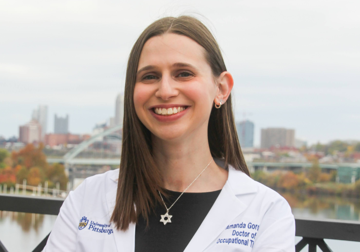 Woman with straight brown hair wearing a white lab coat over a black dress with a silver Star of David necklace and silver earrings.